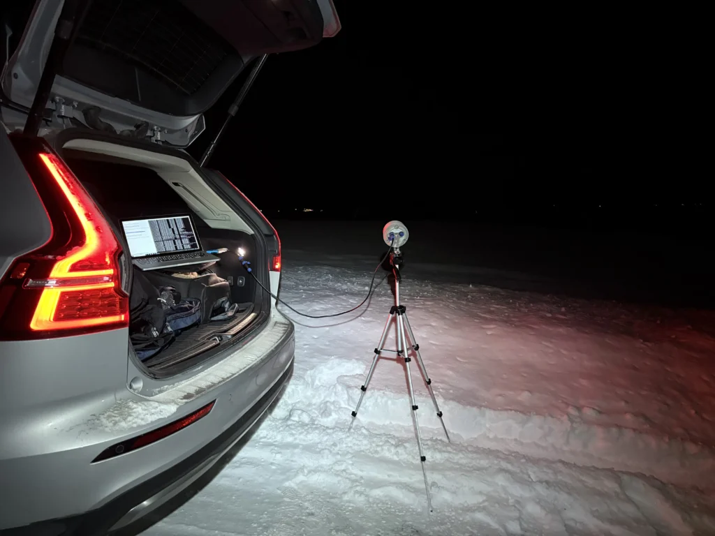 Car trunk open at night in deep snow, revealing a laptop and cables connected to an external satellite antenna on a tripod.