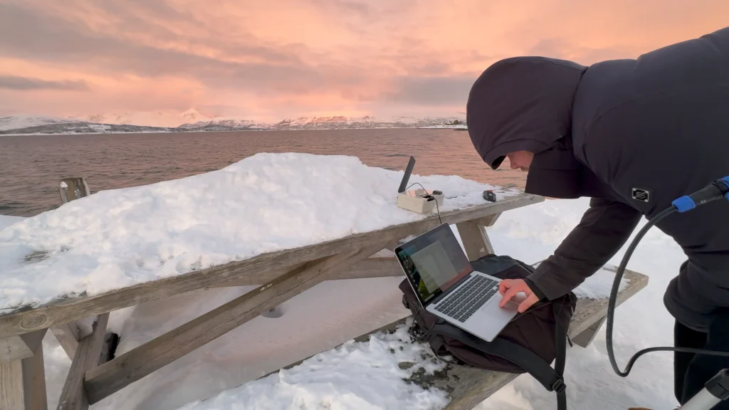 A Tele2 IoT employee in winter gear works on a laptop at a snowy picnic table by a lake with mountains, testing satellite IoT.